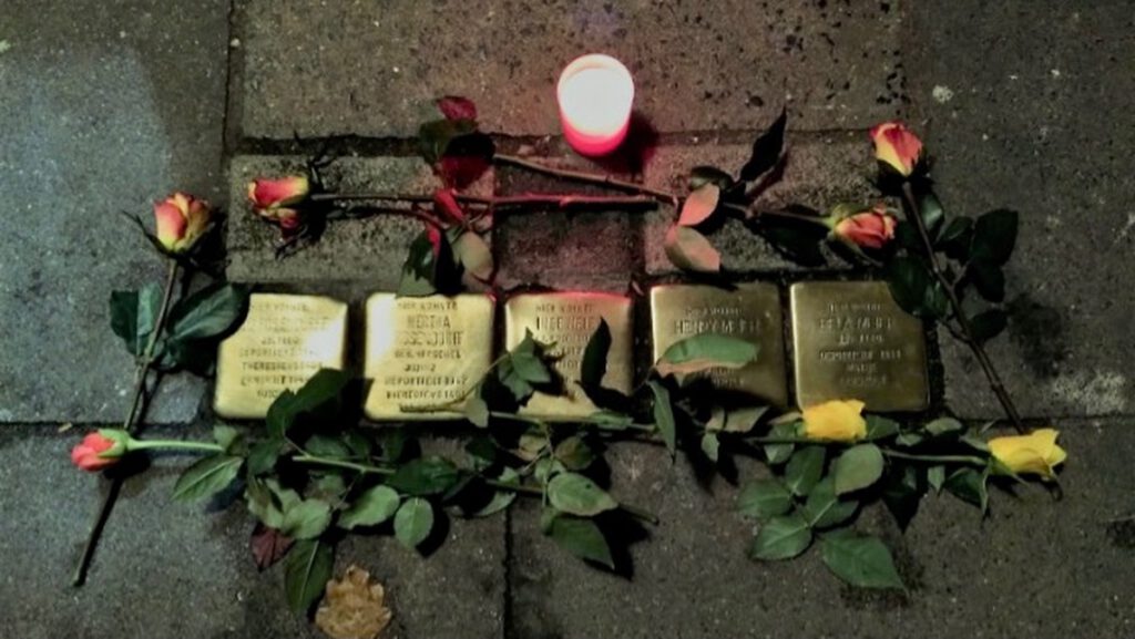 Das Bild zeigt fünf Stolpersteine, die auf fünf deportierte und erforderte Menschen hinweisen, die an diesem Ort gelebt haben. Die Steine sind mit roten und gelben Rosen und einer Kerze versehen. 