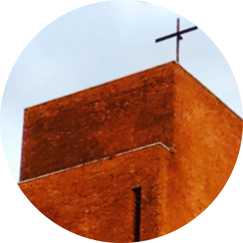 Das Bild zeigt die Turmspitze der Kirche mit einem schlichten Kreuz vor blau-weißem Himmel.