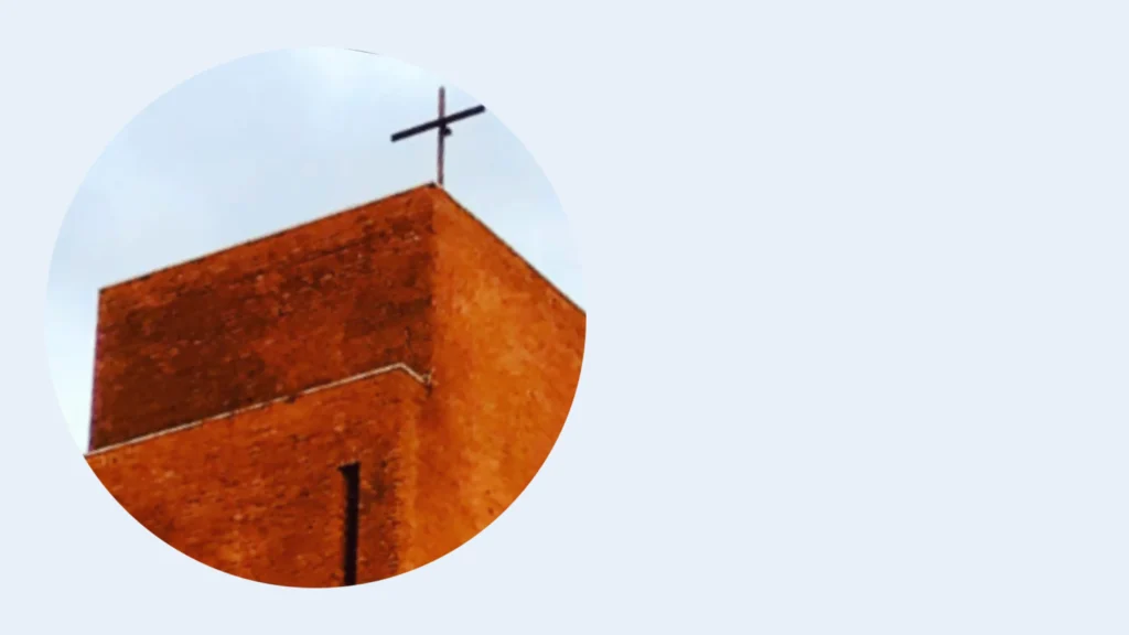 Das Bild zeigt die Turmspitze der Kirche mit einem schlichten Kreuz vor blau-weißem Himmel.